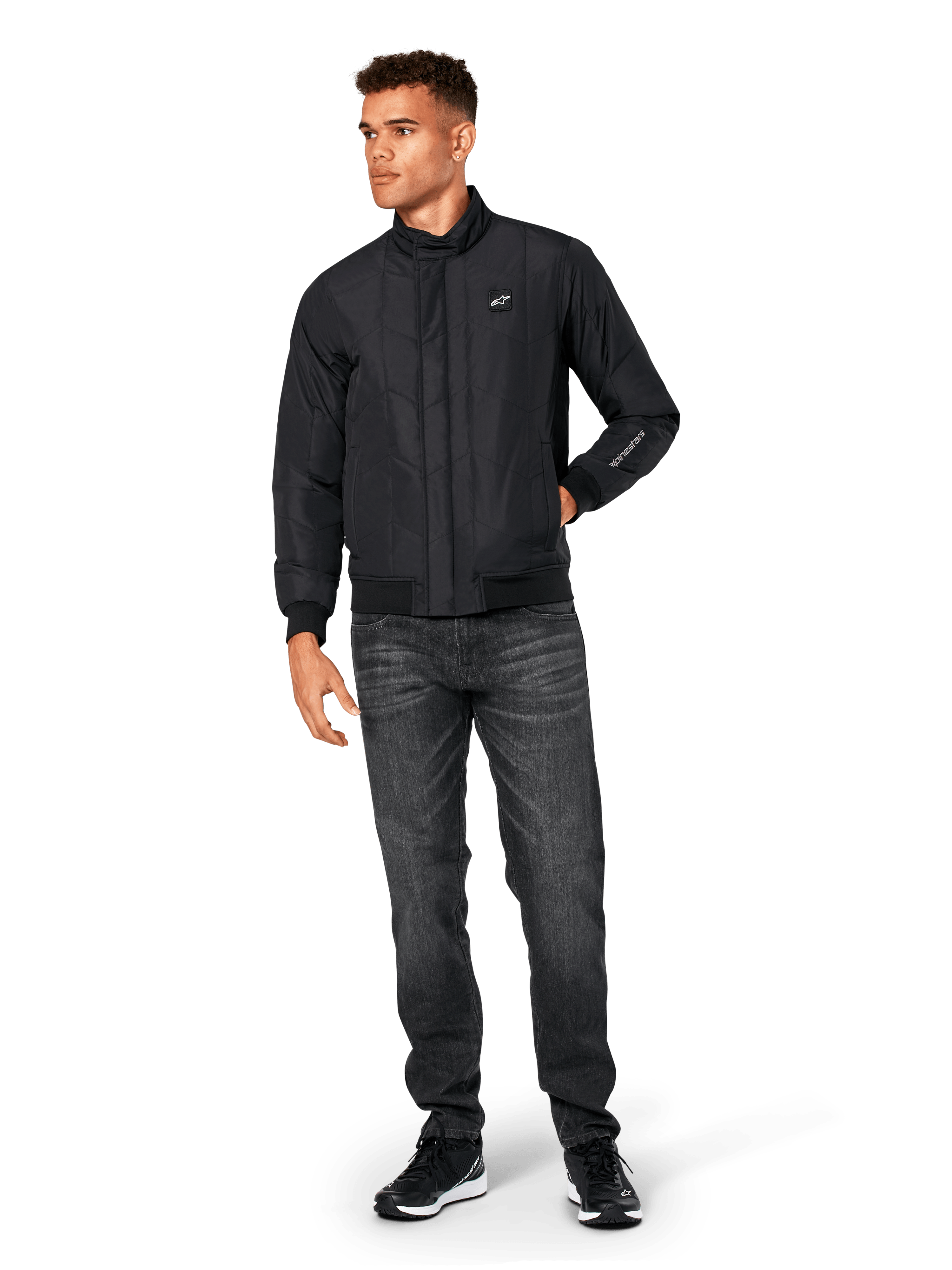 Alpinestars Precedent jas, gesteppte sportswear jas, zwart, gedragen met donkergrijze jeans en zwarte sneakers, voorzien van een schuin gestikt patroon, lage kraag, ribgebreide manchetten en zoom, met een witte logopatch op de borst en borduurwerk op de mouw.