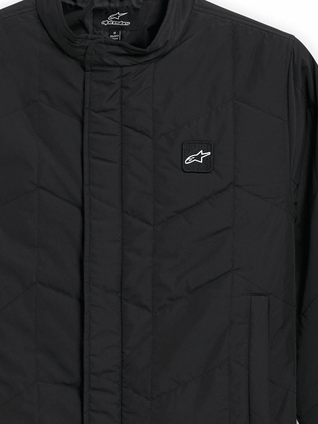 Alpinestars Precedent jas, gestept sportswear jack, zwart, detailopname met een iconisch schuin gestikt patroon, lage kraag, bedekte rits aan de voorzijde en kleine logopatch op de borst