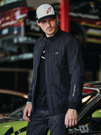 Alpinestars Precedent jas, zwarte gesteppte jas met Formule 1-styling, voorzien van een iconisch schuin gestikt patroon, logopatch op de borst en wit borduurwerk op de mouw, gedragen met een bijpassende Alpinestars pet in een motorsport garageomgeving.