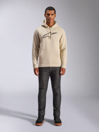 Alpinestars Ageless V3 Hoodie, lässige Sportbekleidung, Off-White mit schwarz gesticktem Logo, getragen von einem Mann mit dunkelgrauer Hose und schwarzen Sneakern, mit Kapuze mit Kordelzug und Kängurutasche, modernes vom Motorsport inspiriertes Design