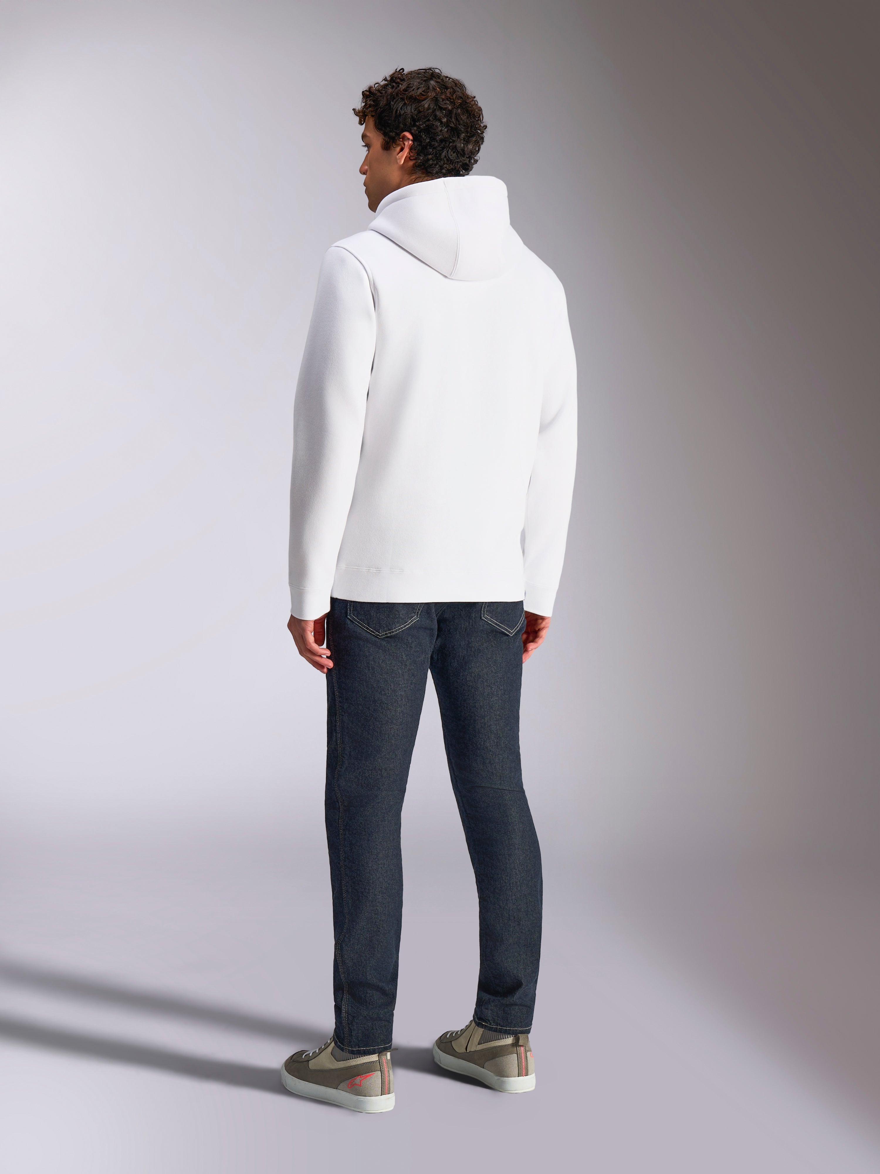 Alpinestars Blaze V3 Hoodie, achteraanzicht van een mannelijk model dat een witte pullover hoodie draagt, casual sportkleding gecombineerd met een donkerblauwe jeans en grijze sneakers, modern op de motorsport geïnspireerd ontwerp