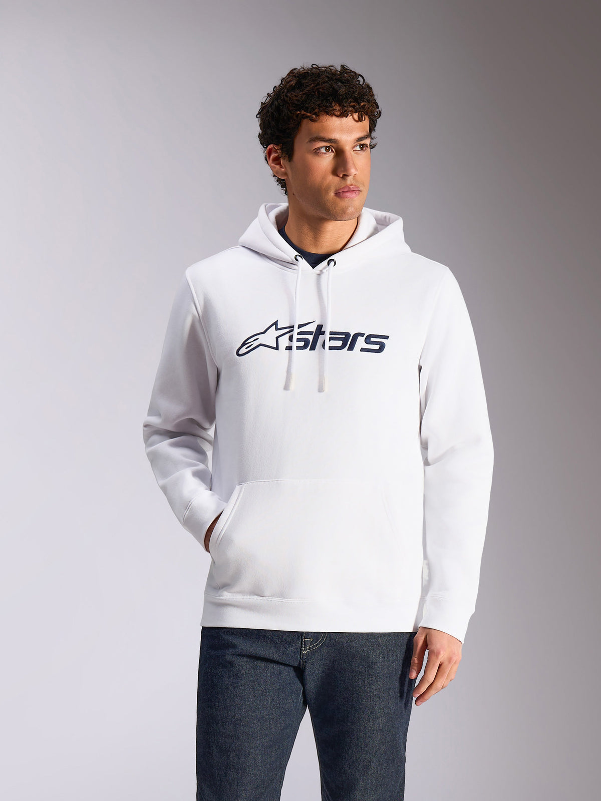 Sweat à capuche Alpinestars Blaze V3, streetwear décontracté, blanc avec logo Alpinestars bleu marine, capuche à cordon de serrage, manches longues, porté avec un jean en denim foncé, design moderne inspiré du sport automobile