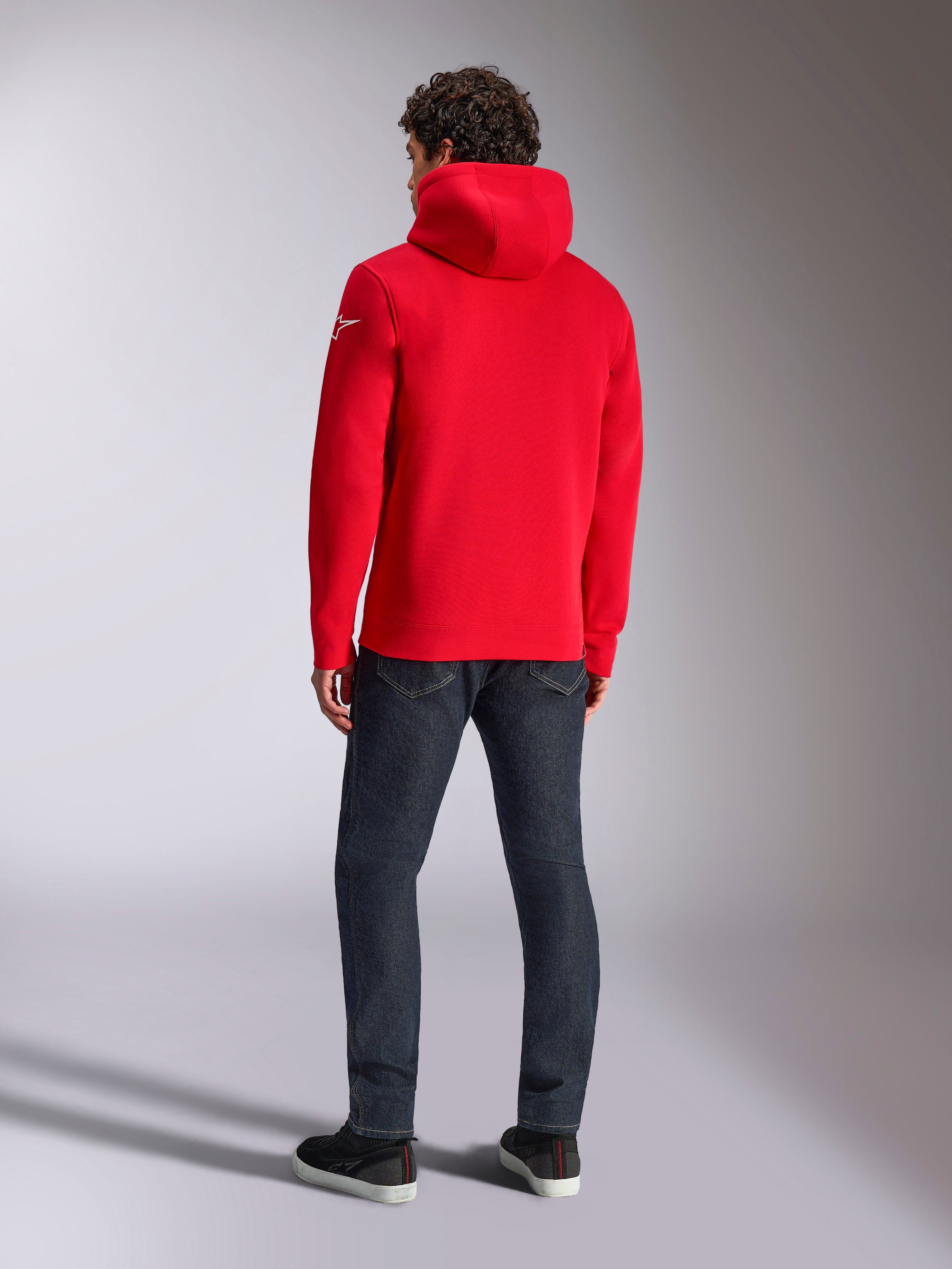Alpinestars Linear Wordmark V3 Hoodie, casual rode pullover hoodie, achteraanzicht met de capuchon en wit Ageless-logo op de mouw, gecombineerd met een donkerblauwe jeans en zwarte schoenen, lifestyle motorsport streetwear
