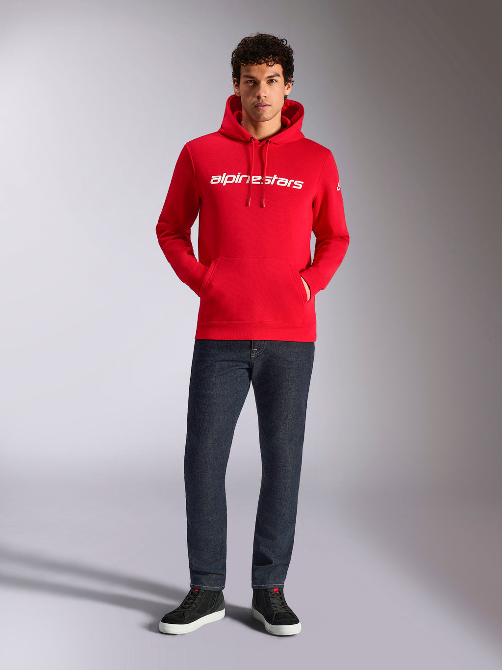 Alpinestars Linear Wordmark V3 Hoodie, casual streetwear, rood met wit lineair woordmerk op de borst, capuchon met trekkoord, kangoeroezak aan de voorzijde, gedragen met donkere jeans en zwarte sneakers, modern op de motorsport geïnspireerd ontwerp