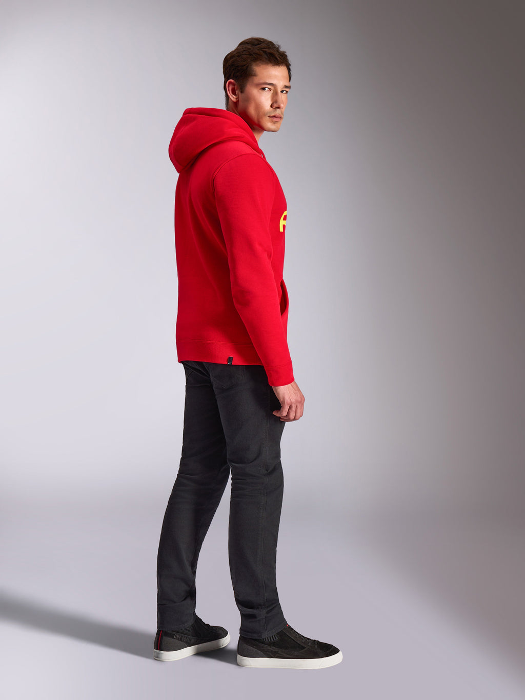 Alpinestars Linear Wordmark V3 Hoodie, rood, casual streetwear, model van de zijkant en achterkant gezien in een rode pullover hoodie met gele logo's, donkergrijze jeans en zwarte sneakers, studio lifestyle-opname