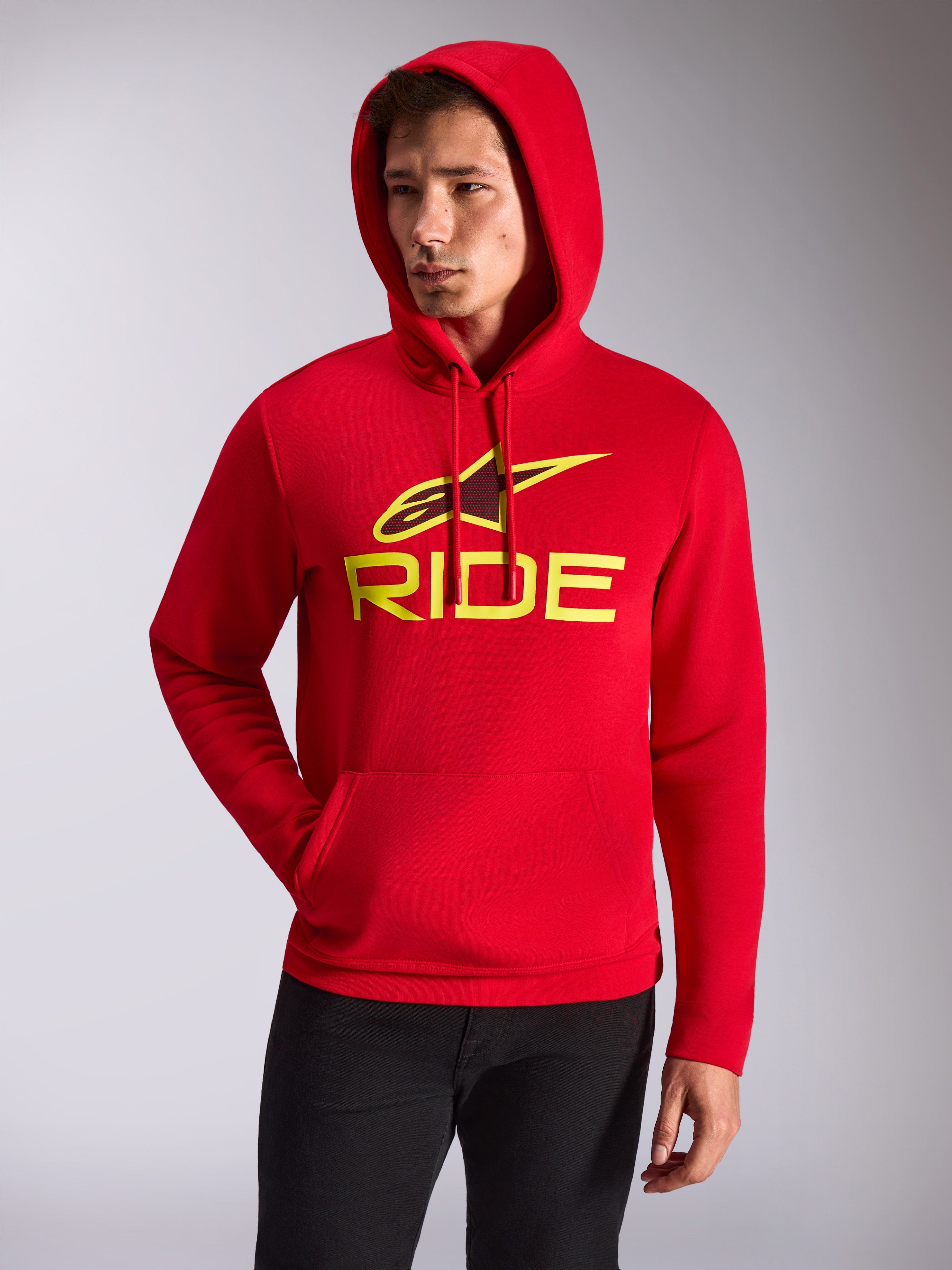 Alpinestars Ride V3 Hoodie, mannelijk model draagt rode pullover fleece met capuchon, hi-vis geel RIDE-logo en Alpinestars-ster op de borst, casual motorsport streetwear, kangoeroezak aan de voorkant, gedragen met zwarte broek