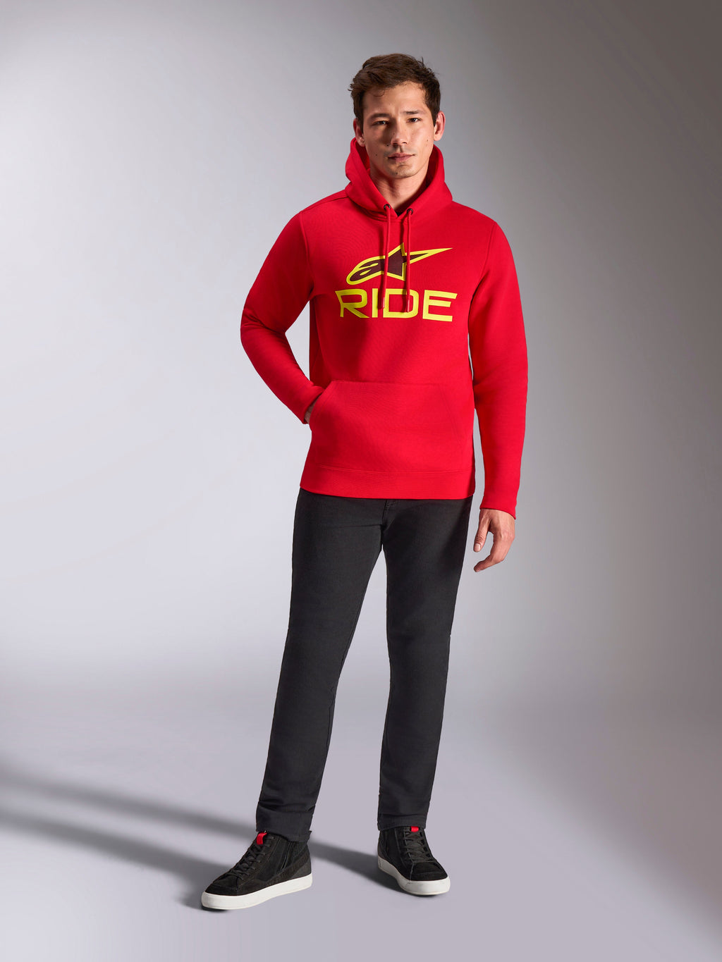 Alpinestars Ride V3 Hoodie, rode fleece hoodie, felgeel RIDE-logo met Astars-symbool op de borst, gedragen met een zwarte broek en zwarte hoge sneakers, casual op motorsport geïnspireerde streetwear