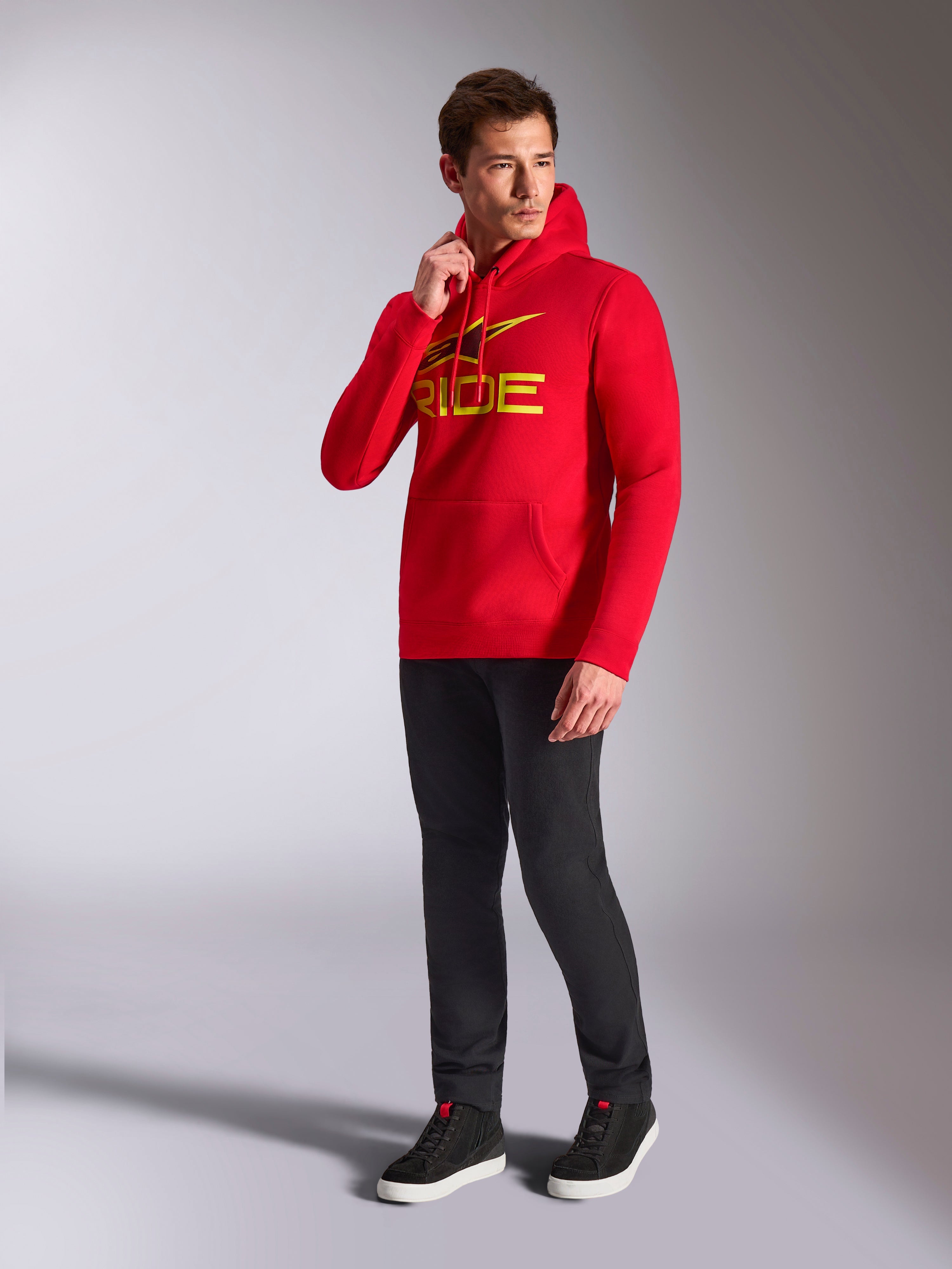 Alpinestars Ride V3 Hoodie, rode pullover sweatshirt, geel en zwart 'RIDE'-logo op de borst, gedragen door model met zwarte broek en zwarte sneakers, casual op motorsport geïnspireerde streetwear
