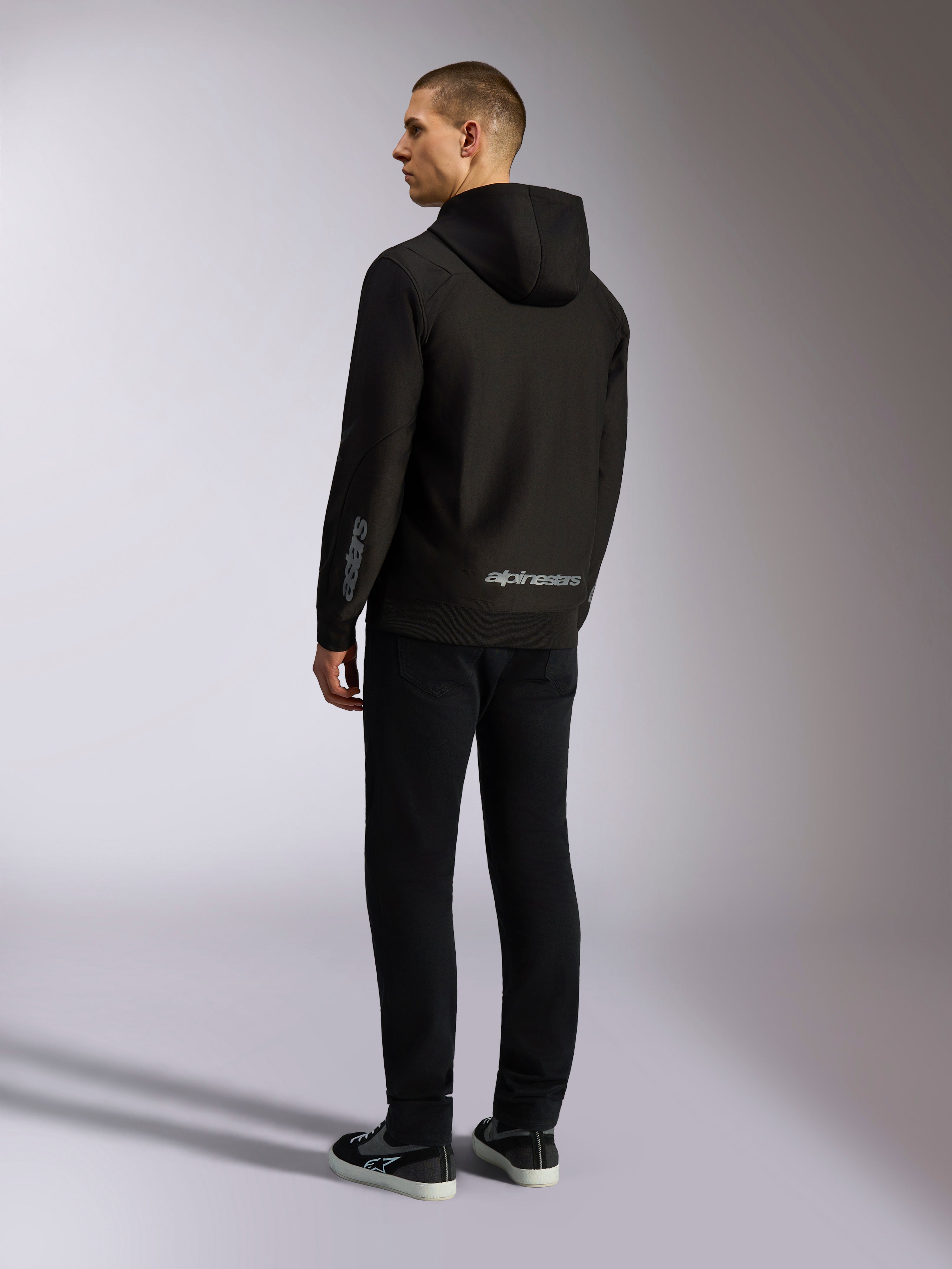 Alpinestars Sessions V3 Hoodie, zwarte casual streetwear, achteraanzicht op een model, met grijs Alpinestars-woordmerk op de onderrug en Astars-logo op de mouw, hoodie met rits aan de voorzijde en moderne motorsport-geïnspireerde styling