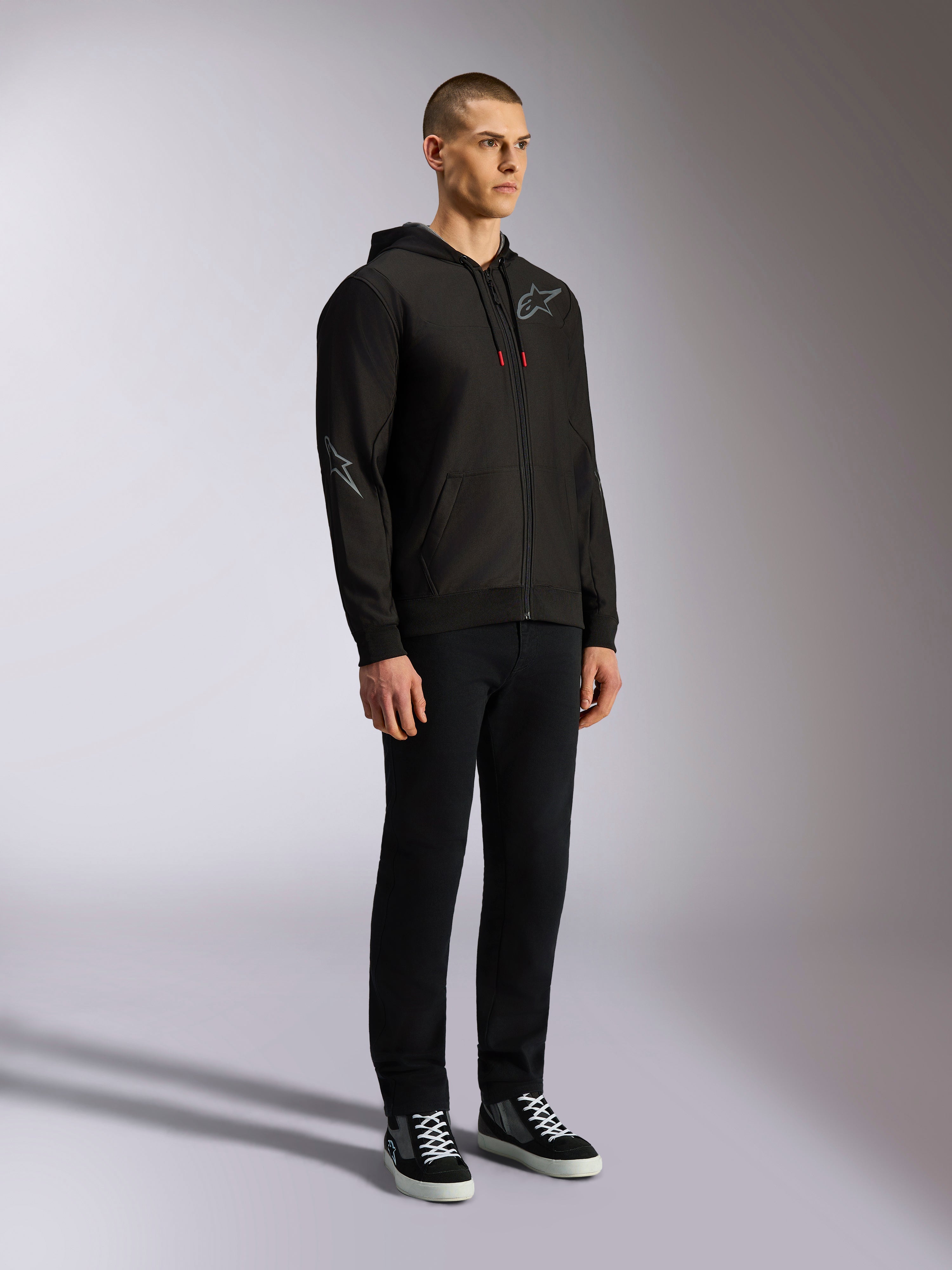 Alpinestars Sessions V3 Hoodie, casual motorsport streetwear, zwarte hoodie met rits en grijs ster-logo op de borst en mouw, trekkoorden met rode uiteinden, geribde manchetten en zoom, gedragen door model met zwarte broek en sneakers.