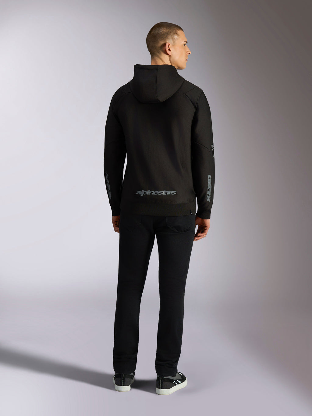 Alpinestars Sessions V3 Hoodie, casual streetwear, zwart met grijs Alpinestars woordmerk op de onderrug en 'astars' branding op de mouwen, achteraanzicht van mannelijk model in hoodie en zwarte broek, modern op de motorsport geïnspireerd ontwerp