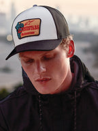 Casquette Trucker Alpinestars Hardware en Military White, portée par un homme en sweat à capuche noir, dotée d'un panneau avant blanc avec visière courbée noire et arrière en maille noire, écusson brodé beige avec marquage Alpinestars Motocross rouge, style lifestyle motorsport streetwear décontracté