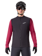 Gilet thermique Alpinestars A-Dura, gilet de cyclisme noir isolé, porté par un mannequin avec un maillot à manches longues bordeaux, casque de VTT noir et lunettes de soleil, avec logo blanc sur la poitrine, protection thermique légère pour le VTT