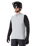 Gilet thermique Alpinestars A-Dura, gris titane, gilet de cyclisme isolant, porté sur un haut noir à manches longues avec un casque de VTT et des gants noirs, arborant un logo rose sur la poitrine et des poches latérales zippées verticales