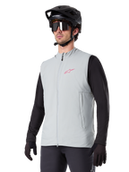Gilet thermique Alpinestars A-Dura, gris titane, gilet de cyclisme isolant, porté sur un haut noir à manches longues avec un casque de VTT et des gants noirs, arborant un logo rose sur la poitrine et des poches latérales zippées verticales
