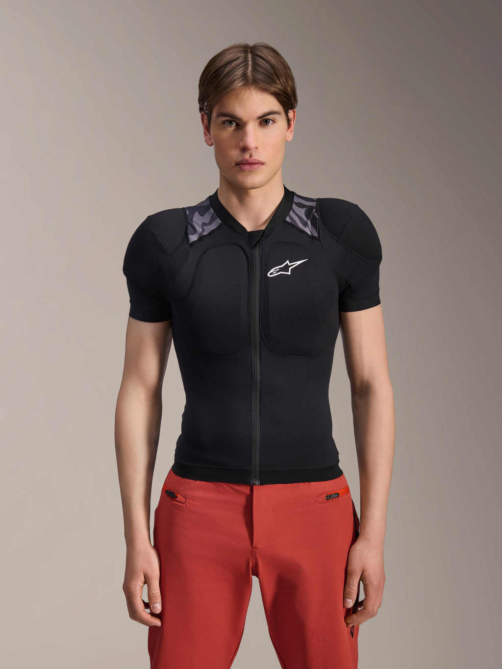 Mannelijk model draagt Alpinestars A-Motion Plasma Protection Jacket - Korte mouwen, zwart met grijze camouflage schoudervulling, wit Alpinestars-logo op de borst, gecombineerd met een rode broek, slim-fit MTB-bodyprotector met een rits aan de voorkant