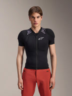 Mannelijk model draagt Alpinestars A-Motion Plasma Protection Jacket - Korte mouwen, zwart met grijze camouflage schoudervulling, wit Alpinestars-logo op de borst, gecombineerd met een rode broek, slim-fit MTB-bodyprotector met een rits aan de voorkant