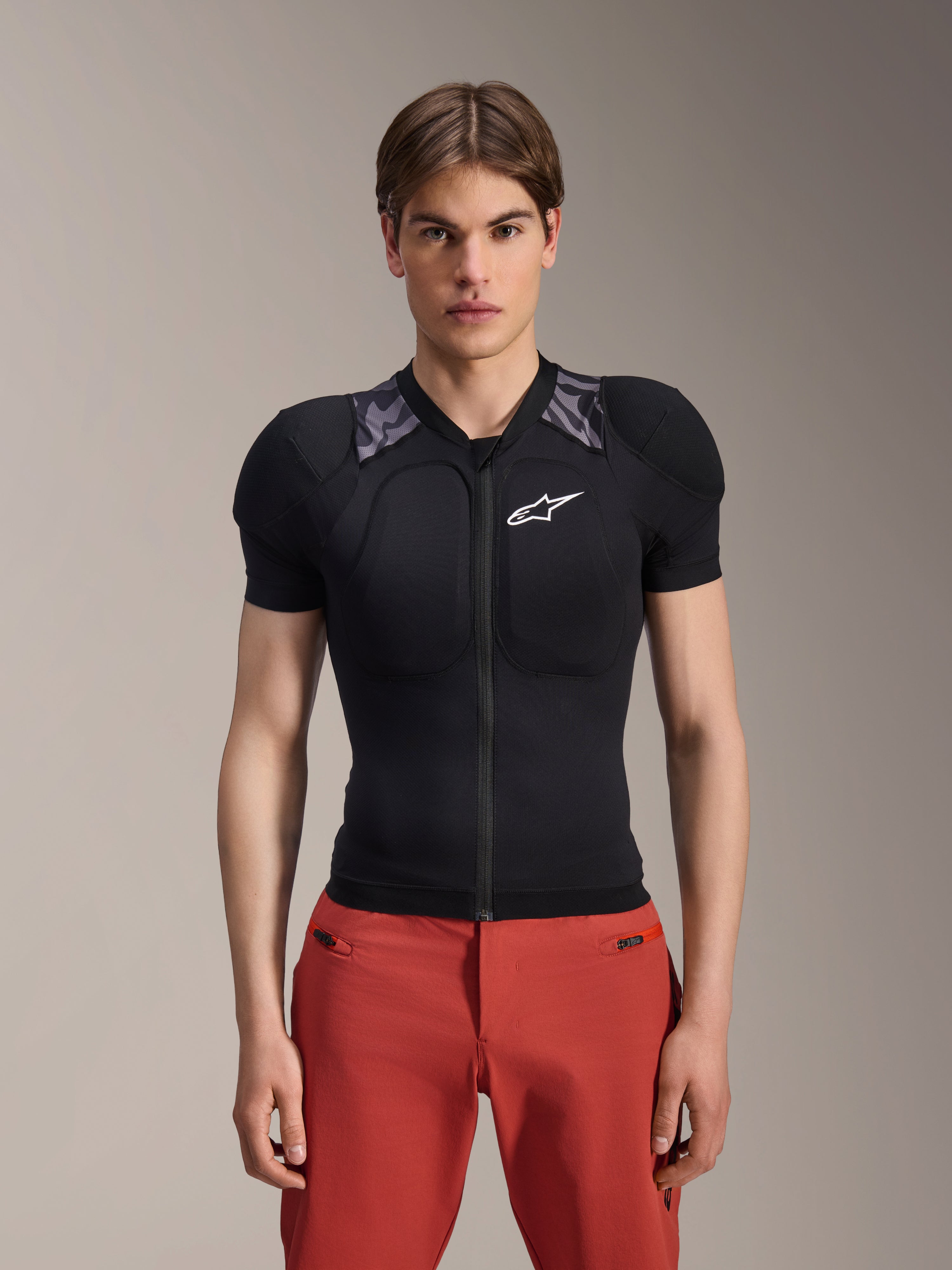 Mannelijk model draagt Alpinestars A-Motion Plasma Protection Jacket - Korte mouwen, zwart met grijze camouflage schoudervulling, wit Alpinestars-logo op de borst, gecombineerd met een rode broek, slim-fit MTB-bodyprotector met een rits aan de voorkant