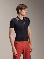 Mannelijk model draagt Alpinestars A-Motion Plasma beschermjas met korte mouwen, Black Camo met grijze camouflage schouderpanelen, voorzien van geïntegreerde schouder- en borstbeschermers, wit ster-logo op de borst, ademende mesh-stof, gecombineerd met een rode broek