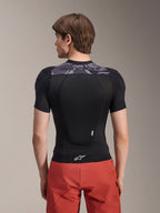Alpinestars A-Motion Plasma Protection Jacket - Korte Mouwen, zwart met grijze camo-accenten op de bovenrug, achteraanzicht met geïntegreerde rugprotector en wit Astars-logo op de zoom, gedragen met rode short, technische mountainbike-beschermingsuitrusting