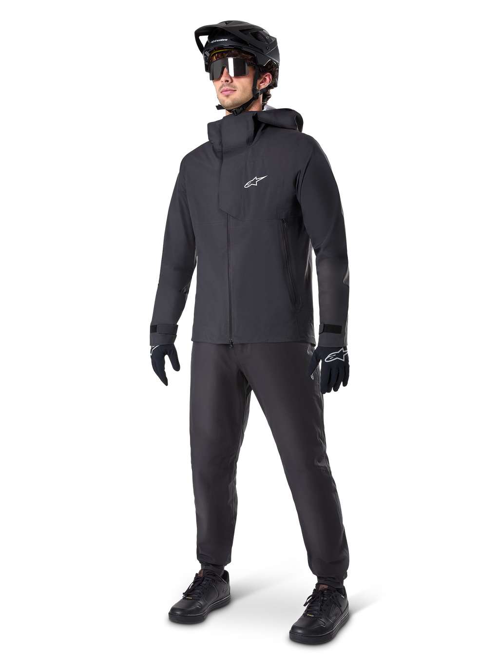Vététiste portant le pantalon imperméable Alpinestars A-Dura noir, associé à une veste imperméable à capuche noire assortie, un casque de VTT noir avec visière, des lunettes de cyclisme teintées et des gants noirs, équipement professionnel complet pour le trail par tous les temps
