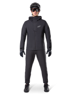 Pantalon imperméable Alpinestars A-Dura, pantalon de VTT noir, porté avec un équipement de VTT complet noir incluant une veste technique imperméable, un casque noir, des lunettes de soleil et des gants, conçu pour le tout-terrain par temps humide.