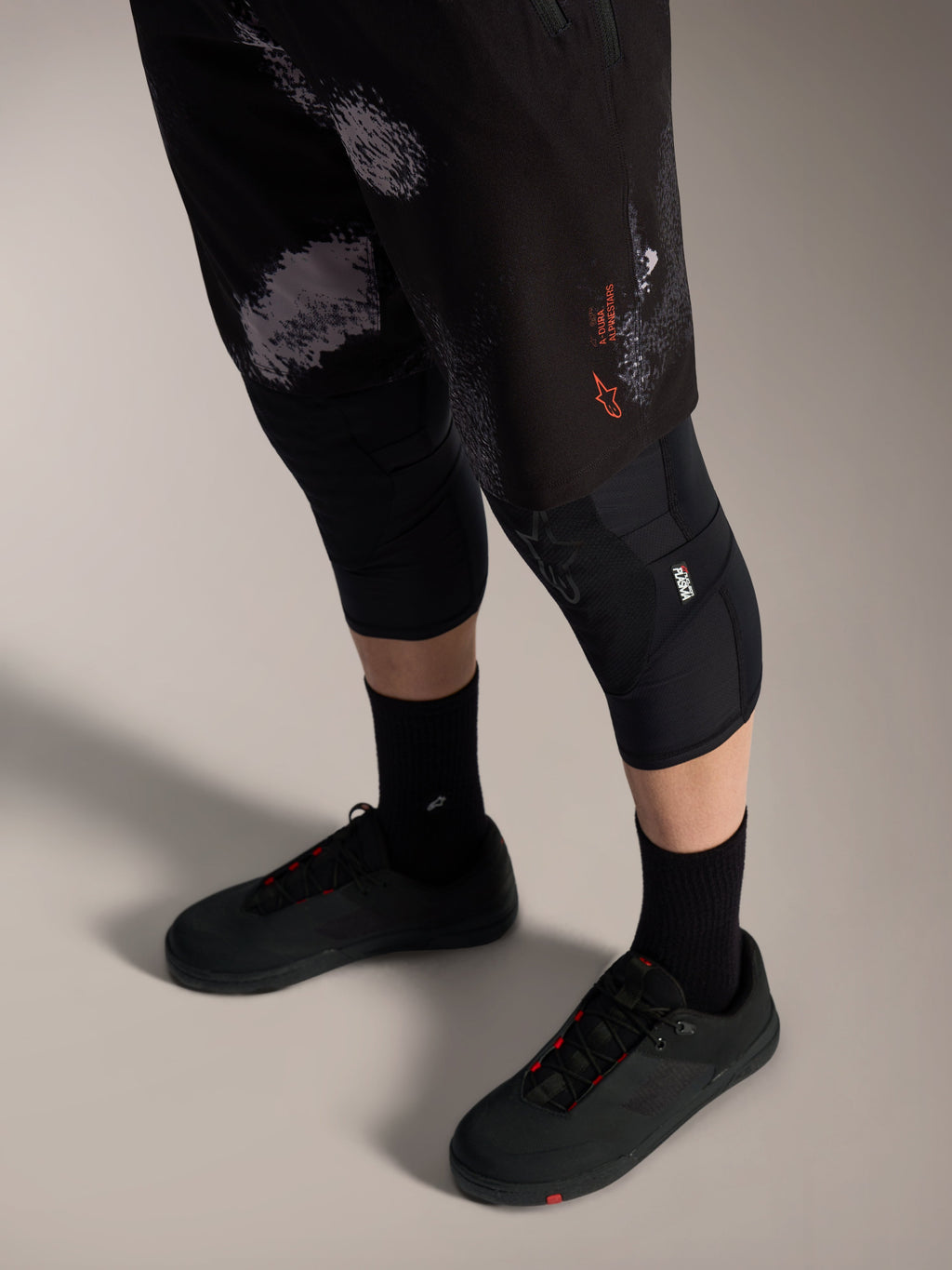 Alpinestars A-Dura Ride MTB-short in Black Camo met oranje logo, gedragen met kniebeschermers en fietsschoenen.