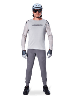 Pantalon Alpinestars A-Aria Elite, pantalon de VTT gris foncé, porté par un cycliste en maillot à manches longues gris clair, casque bleu clair et chaussures noires, doté de perforations laser visibles à l'intérieur des jambes et d'une coupe performance ajustée.