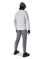 Pantalon Alpinestars A-Aria Elite, pantalon de cyclisme VTT, gris foncé, vue arrière d'un cycliste portant une tenue complète, logos Alpinestars blancs sur le bas des jambes, coupe performance avec perforations laser, associé à un maillot à manches longues gris clair et un casque de VTT gris