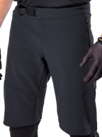 Alpinestars A-Aria Elite Shorts in zwart, close-up van de taille en dij, voorzien van magnetische gespsluiting, zakken met ritssluiting en lasergesneden ventilatieperforaties, gedragen met zwarte fietshandschoenen voor mountainbiken