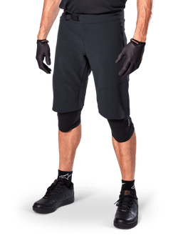 Alpinestars A-Aria Elite Shorts, short de cyclisme noir, porté avec des gants et des genouillères noirs assortis, doté d'une boucle magnétique et de perforations découpées au laser, design épuré pour le VTT tout-terrain