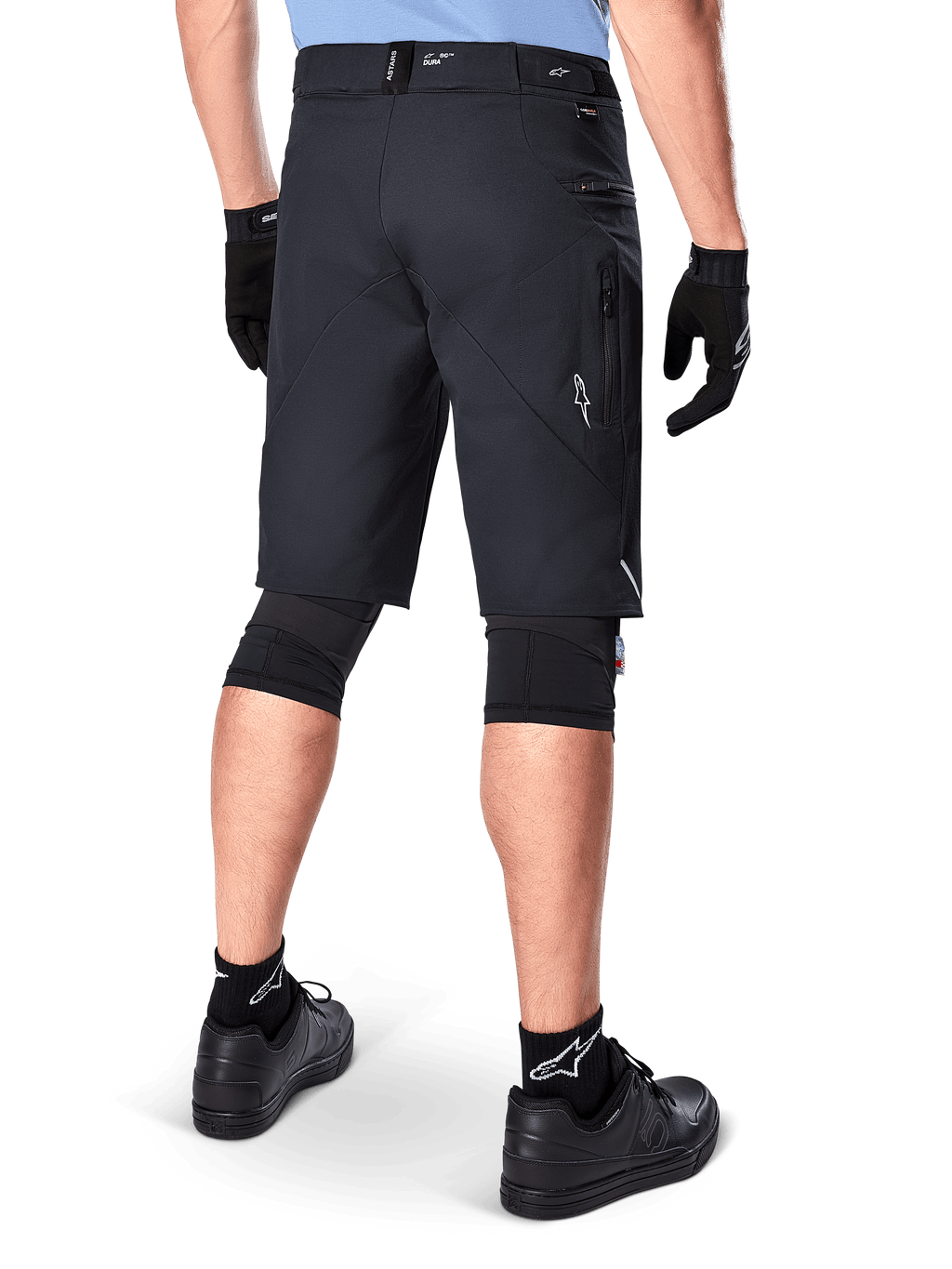 Shorts Alpinestars A-Dura Elite, Noir, vue arrière d'un cycliste portant un équipement de VTT comprenant des genouillères, des gants et des chaussettes noirs avec des logos blancs, doté de poches latérales zippées et d'accents réfléchissants sur un tissu Cordura durable.