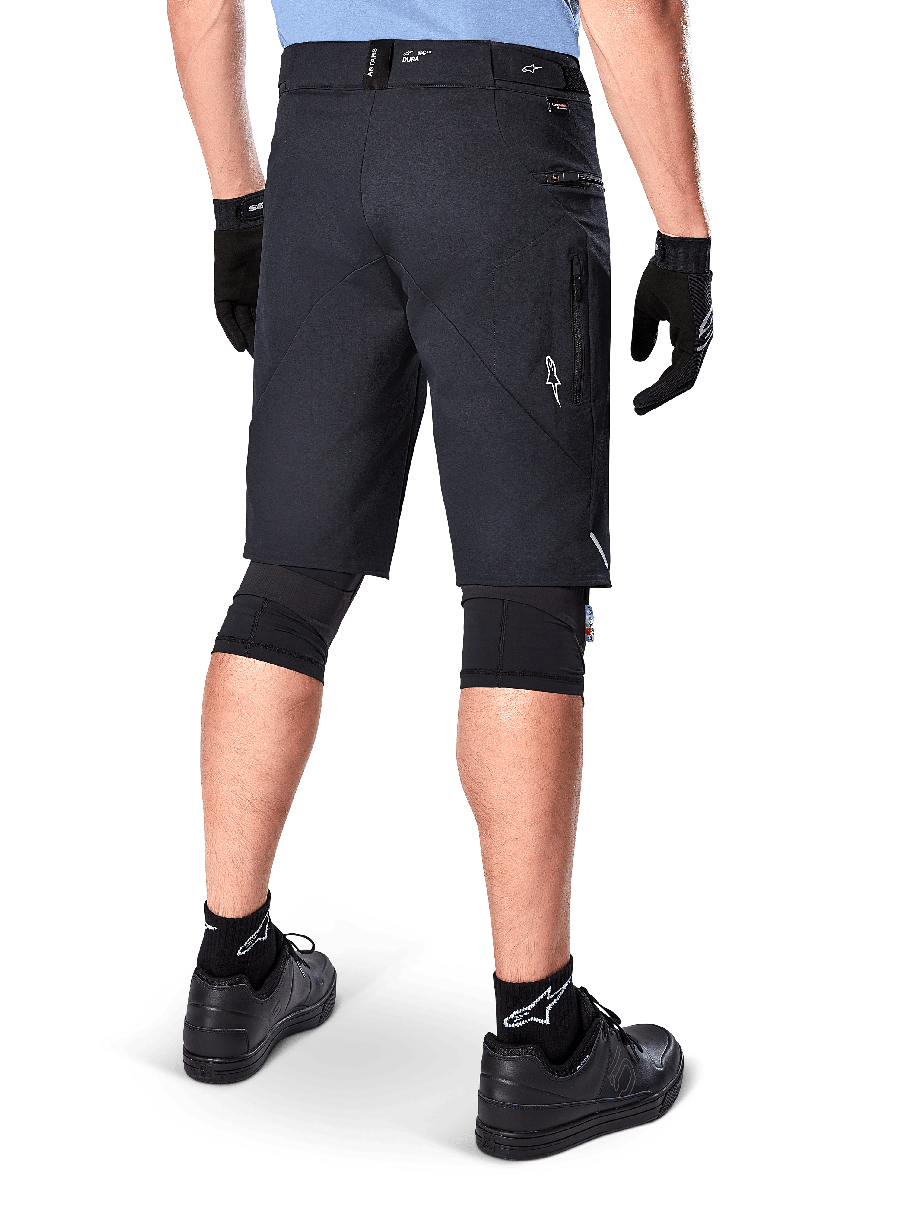 Shorts Alpinestars A-Dura Elite, Noir, vue arrière d'un cycliste portant un équipement de VTT comprenant des genouillères, des gants et des chaussettes noirs avec des logos blancs, doté de poches latérales zippées et d'accents réfléchissants sur un tissu Cordura durable.