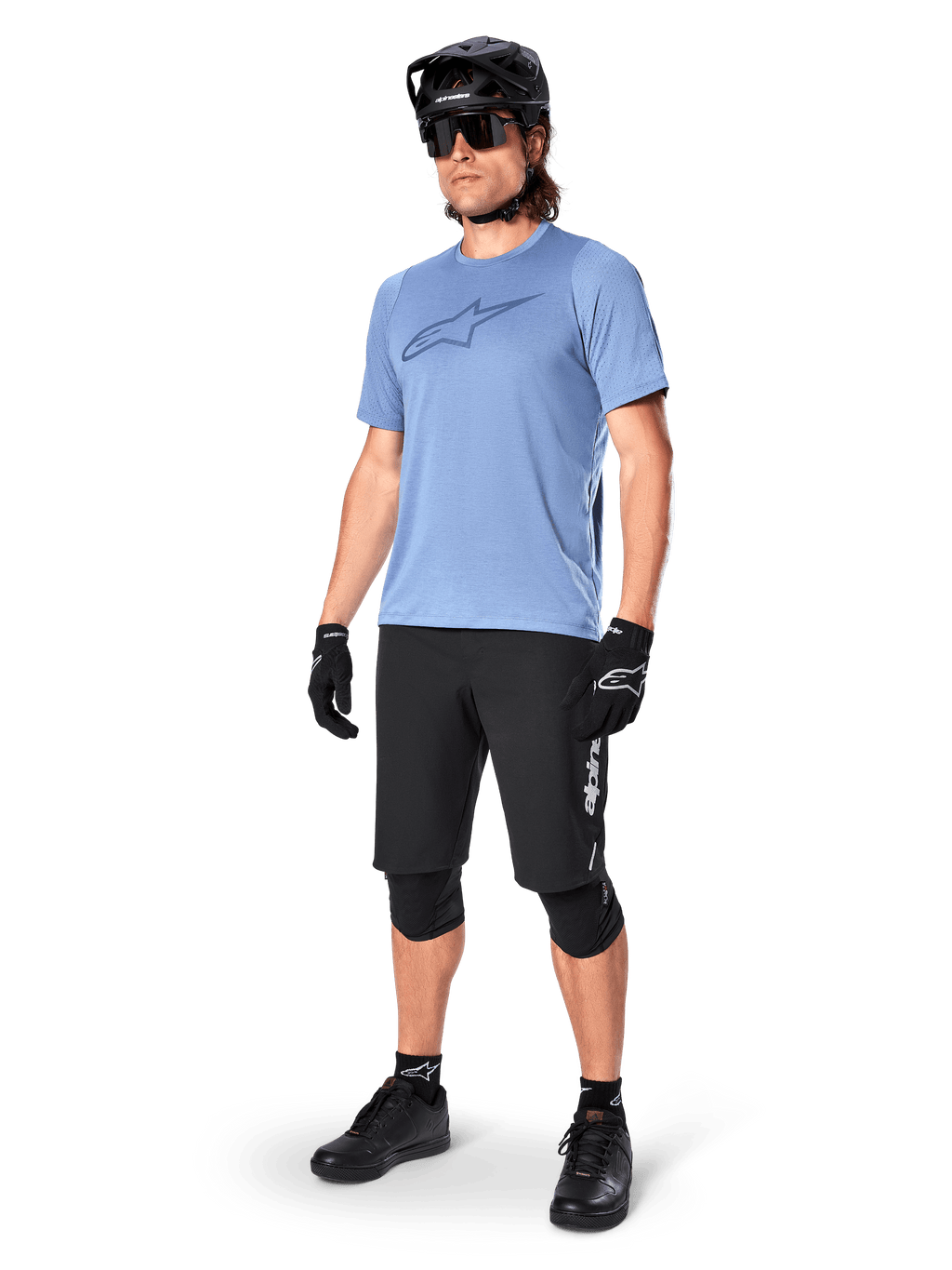 Cycliste portant le short Alpinestars A-Dura Elite noir, maillot bleu clair à manches courtes avec logo sur la poitrine, casque noir, lunettes de soleil et genouillères de protection, équipement complet de VTT pour les sentiers tout-terrain.