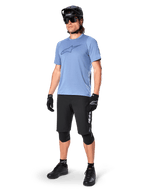 Cycliste portant le short Alpinestars A-Dura Elite noir, maillot bleu clair à manches courtes avec logo sur la poitrine, casque noir, lunettes de soleil et genouillères de protection, équipement complet de VTT pour les sentiers tout-terrain.
