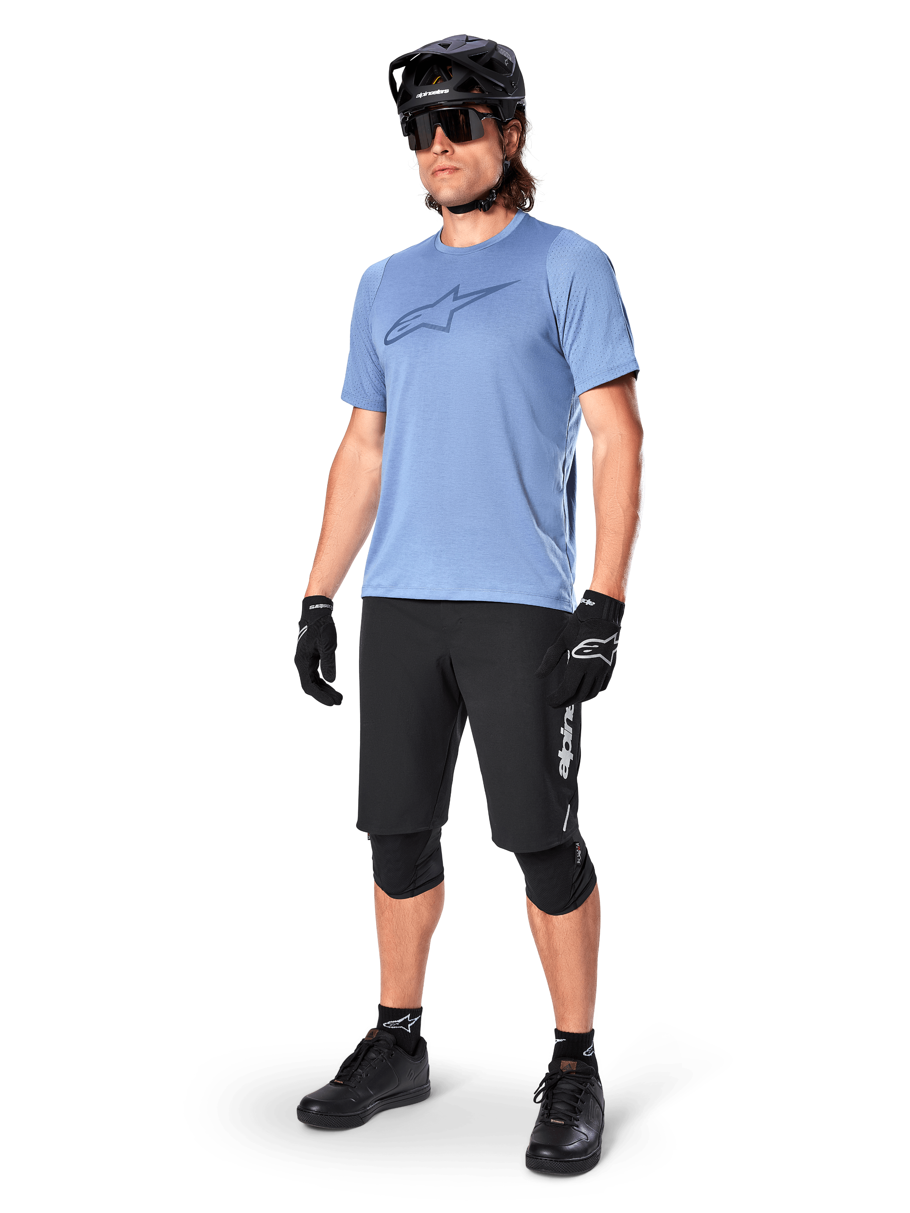 Cycliste portant le short Alpinestars A-Dura Elite noir, maillot bleu clair à manches courtes avec logo sur la poitrine, casque noir, lunettes de soleil et genouillères de protection, équipement complet de VTT pour les sentiers tout-terrain.
