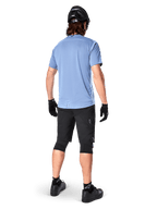 Short Alpinestars A-Dura Elite, short de VTT, noir, vue arrière sur modèle, porté avec un t-shirt bleu clair à manches courtes et équipement de protection noir, design technique avec poches zippées et détails réfléchissants pour le cyclisme tout-terrain