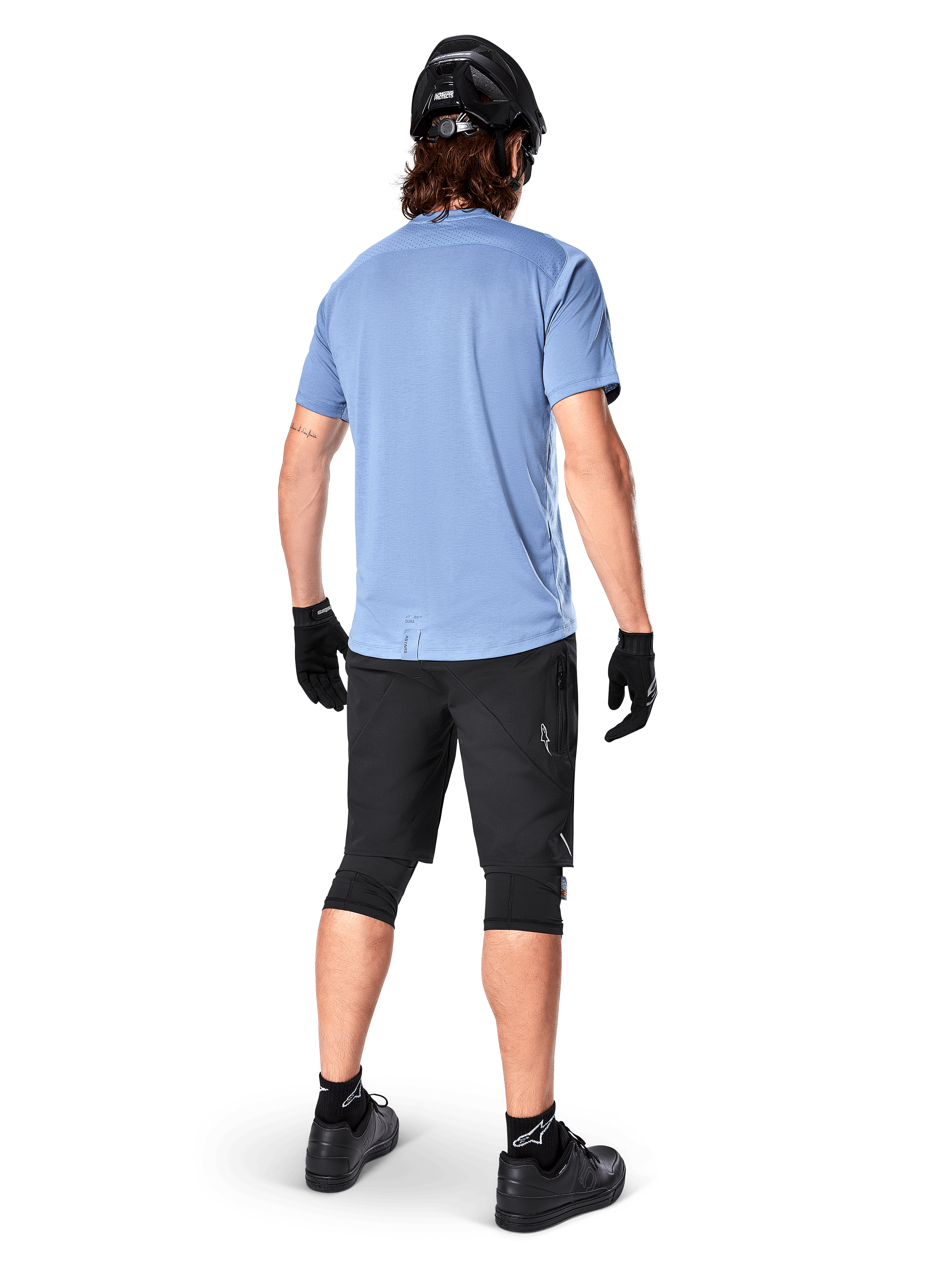 Short Alpinestars A-Dura Elite, short de VTT, noir, vue arrière sur modèle, porté avec un t-shirt bleu clair à manches courtes et équipement de protection noir, design technique avec poches zippées et détails réfléchissants pour le cyclisme tout-terrain