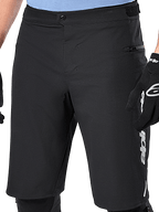 Short Alpinestars A-Dura Elite, short de VTT noir, vue rapprochée présentant la fermeture à bouton-pression à l'avant et une poche latérale zippée, porté par un cycliste avec gants de vélo noirs et genouillères, équipement de performance pour le VTT