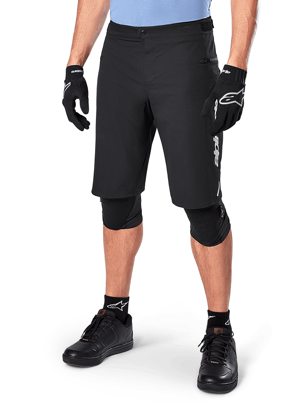 Short Alpinestars A-Dura Elite, short de VTT noir, porté avec un maillot bleu clair et des protections de genoux noires, doté d'une fermeture à bouton-pression sécurisée et d'une poche zippée, associé à des chaussures de cyclisme et des gants noirs pour une tenue tout-terrain complète.