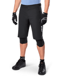 Short Alpinestars A-Dura Elite, short de VTT noir, porté avec un maillot bleu clair et des protections de genoux noires, doté d'une fermeture à bouton-pression sécurisée et d'une poche zippée, associé à des chaussures de cyclisme et des gants noirs pour une tenue tout-terrain complète.
