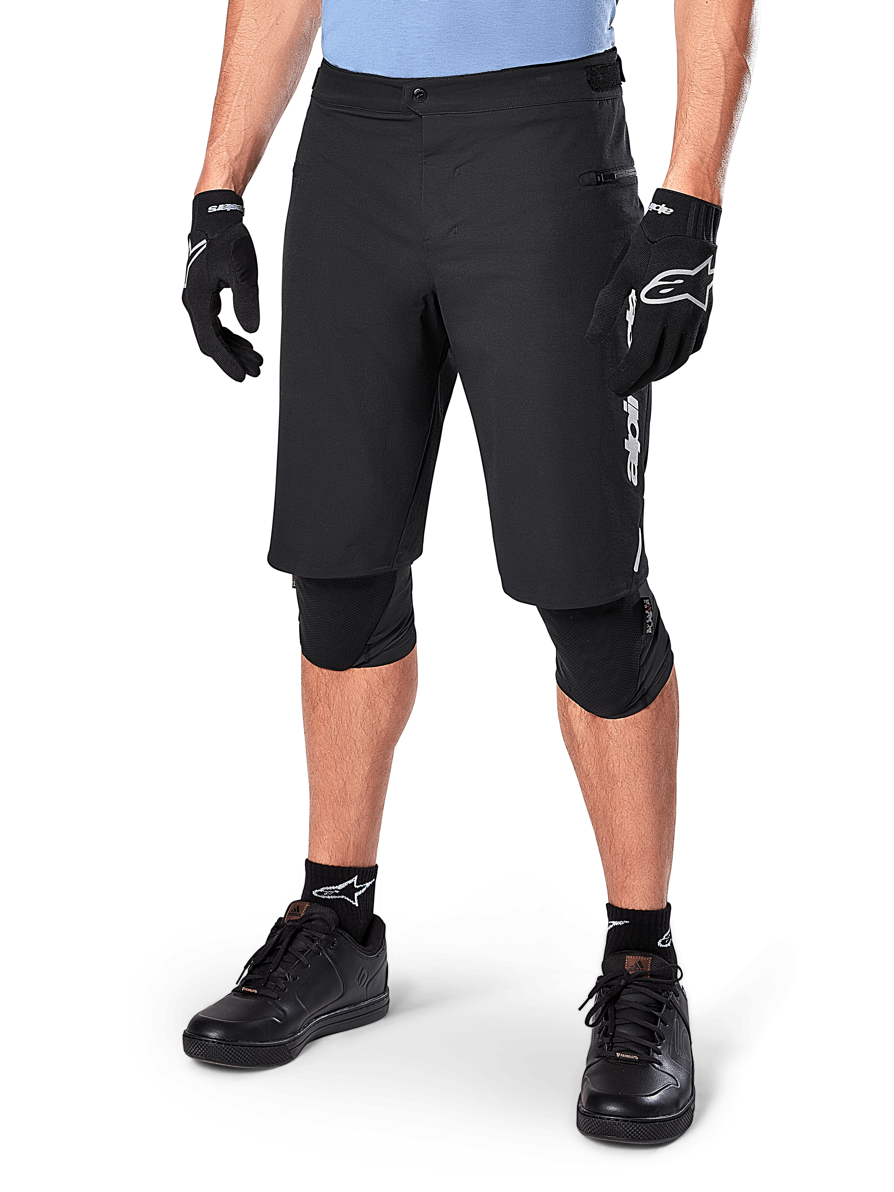 Short Alpinestars A-Dura Elite, short de VTT noir, porté avec un maillot bleu clair et des protections de genoux noires, doté d'une fermeture à bouton-pression sécurisée et d'une poche zippée, associé à des chaussures de cyclisme et des gants noirs pour une tenue tout-terrain complète.