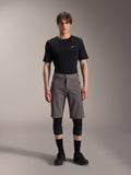 Alpinestars A-Dura Elite Shorts, dunkelgraue Mountainbike-Shorts, getragen mit einem schwarzen T-Shirt mit Rundhalsausschnitt und schwarzen Knieschützern, knielange Performance-Radsportbekleidung mit Druckknopfverschluss vorne und strapazierfähigem Cordura-Gewebe.
