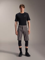 Alpinestars A-Dura Elite Shorts, dunkelgraue Mountainbike-Shorts, getragen mit einem schwarzen T-Shirt mit Rundhalsausschnitt und schwarzen Knieschützern, knielange Performance-Radsportbekleidung mit Druckknopfverschluss vorne und strapazierfähigem Cordura-Gewebe.
