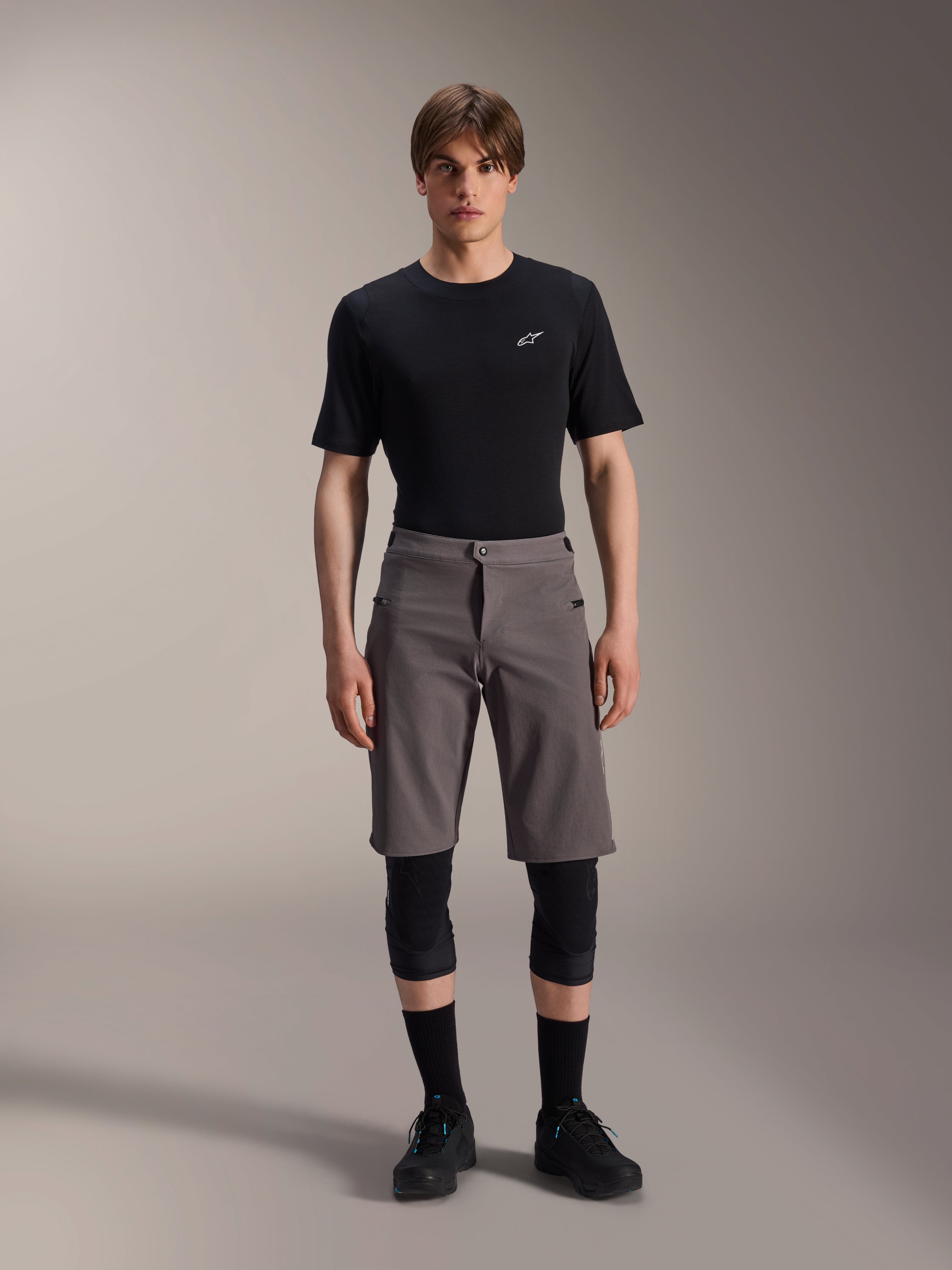 Alpinestars A-Dura Elite Shorts, Dark Gray Mountain Bike Shorts, worn with a black crew neck t-shirt and black knee guards, knee-length performance cycling apparel featuring a front snap closure and durable Cordura fabric.