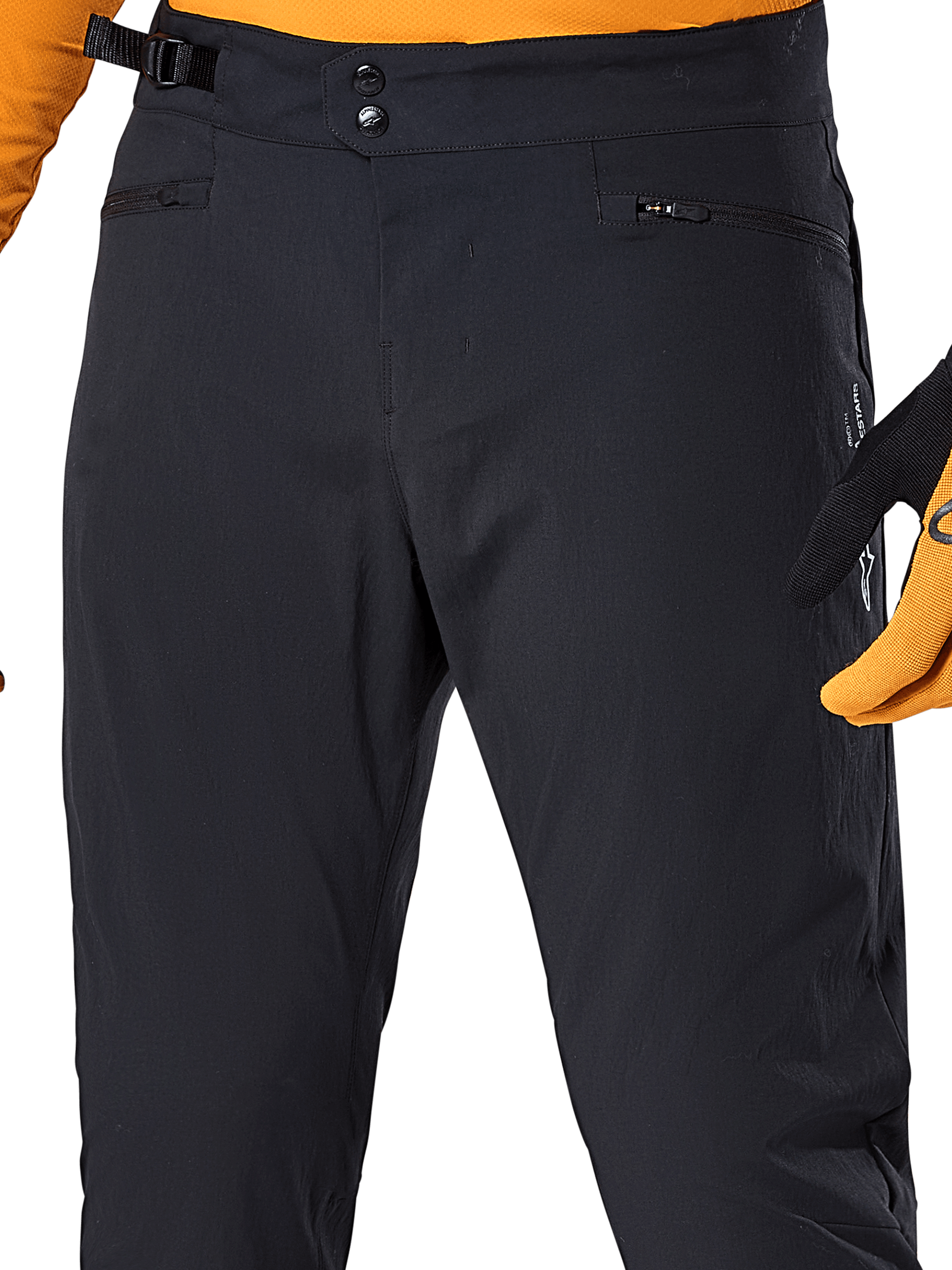 Alpinestars A-Dura Pants, zwarte MTB-broek, close-up met dubbele drukknoopsluiting, verstelbare tailleband aan de zijkant en zakken met rits, gecombineerd met een oranje jersey en handschoenen