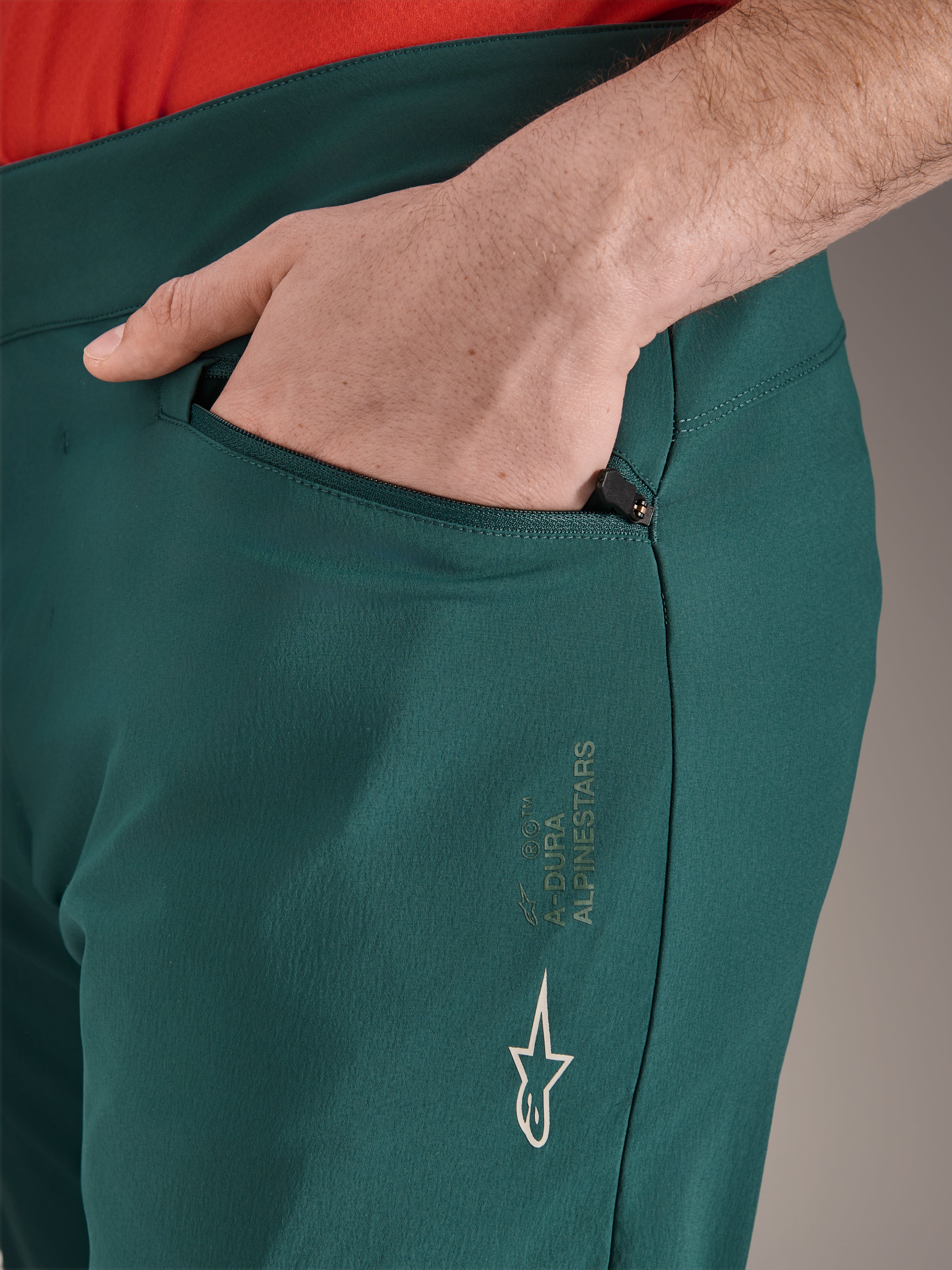 Alpinestars A-Dura Hose in Dark Pine Grün, Nahaufnahme einer Reißverschlusstasche mit Hand, weißes Alpinestars-Logo und Markenschriftzug am Oberschenkel, strapazierfähige MTB-Fahrradhose