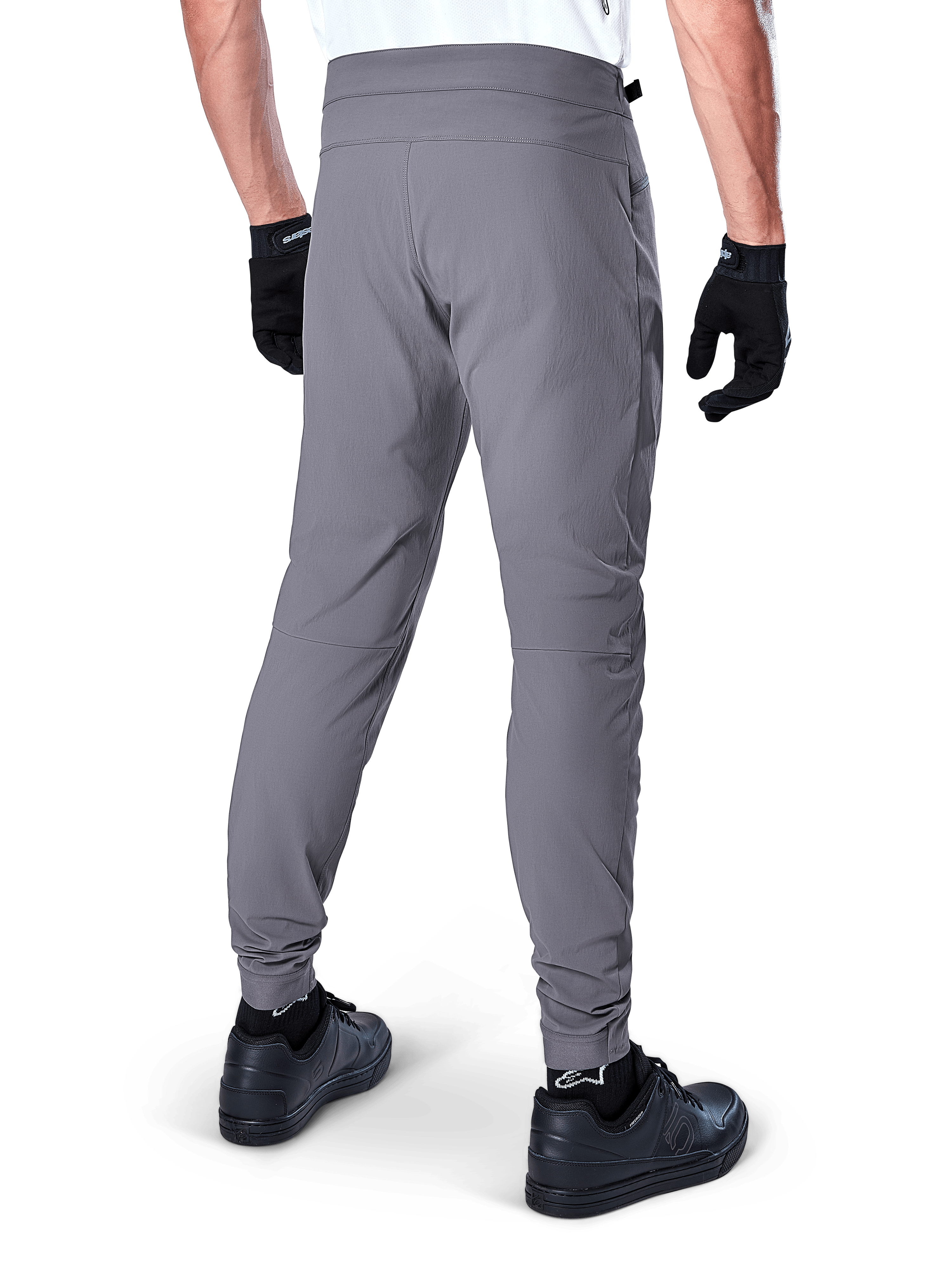 Alpinestars A-Dura Hose, Mountainbike-Hose, Dunkelgrau, Rückansicht eines Modells beim Tragen der Ausrüstung, mit strapazierfähigem 2-Wege-Stretch-Material und elastischen Bündchen, kombiniert mit schwarzen Handschuhen und Sneakern für einen radsporttauglichen Look