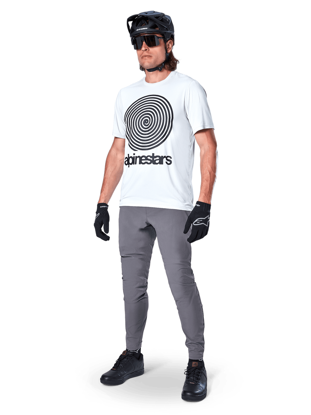 Alpinestars A-Dura Hose, MTB-Hose, Dunkelgrau, getragen mit weißem T-Shirt mit Spiralgrafik, schwarzem MTB-Helm, Sonnenbrille und schwarzen Handschuhen, strapazierfähiges 2-Wege-Stretch-Textil für den Offroad-Radsport