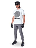 Alpinestars A-Dura Hose, MTB-Hose, Dunkelgrau, getragen mit weißem T-Shirt mit Spiralgrafik, schwarzem MTB-Helm, Sonnenbrille und schwarzen Handschuhen, strapazierfähiges 2-Wege-Stretch-Textil für den Offroad-Radsport